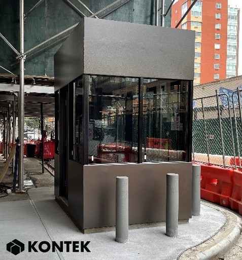 A guard booth installed on a sidewalk, surrounded by bollards for protection. The booth has large ballistic glass windows and a sturdy, modern design. 