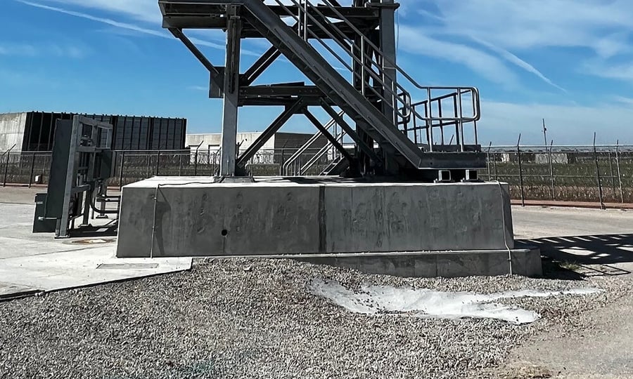 Close-up view of a pre-cast above-grade concrete foundation supporting a ballistic guard tower, with attached metal stairs and a gravel-covered ground in the foreground.