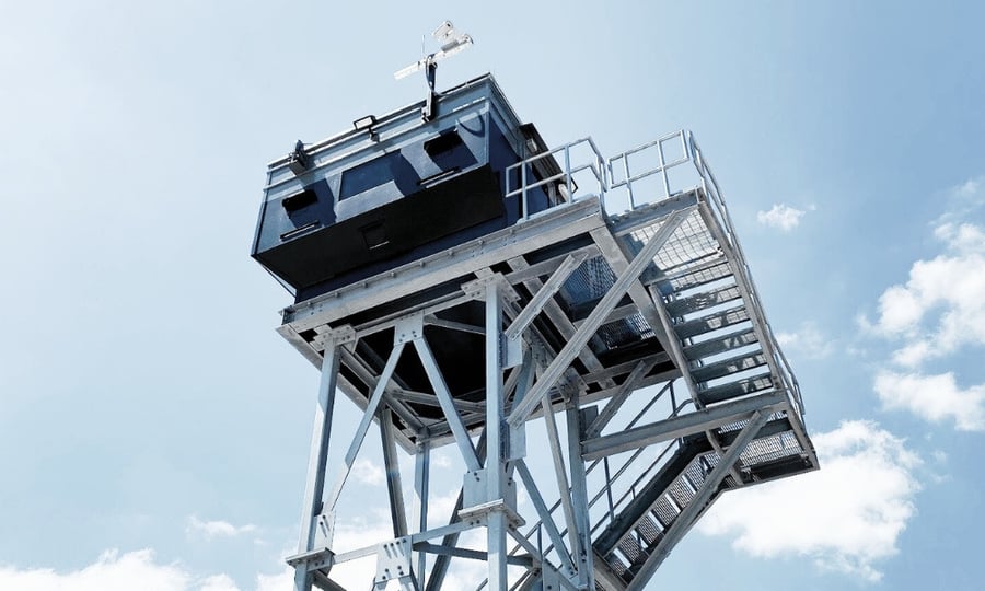 Upward view of an elevated guard tower with a reinforced steel structure, secure guard booth, and surveillance camera mounted on top.